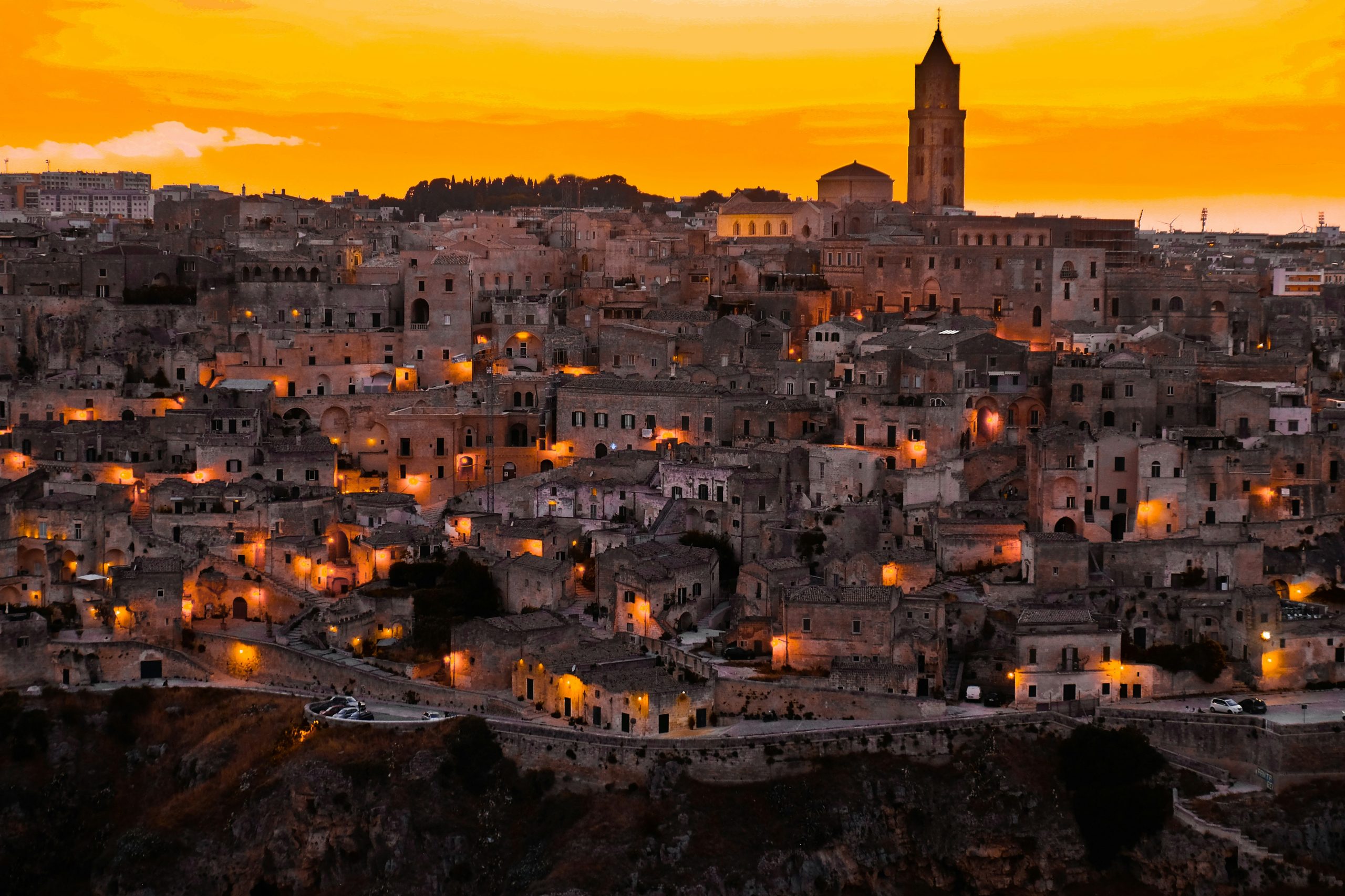 The Sassi of Matera glowing under an orange sunset sky, the cathedral bell tower rising above the old stone town