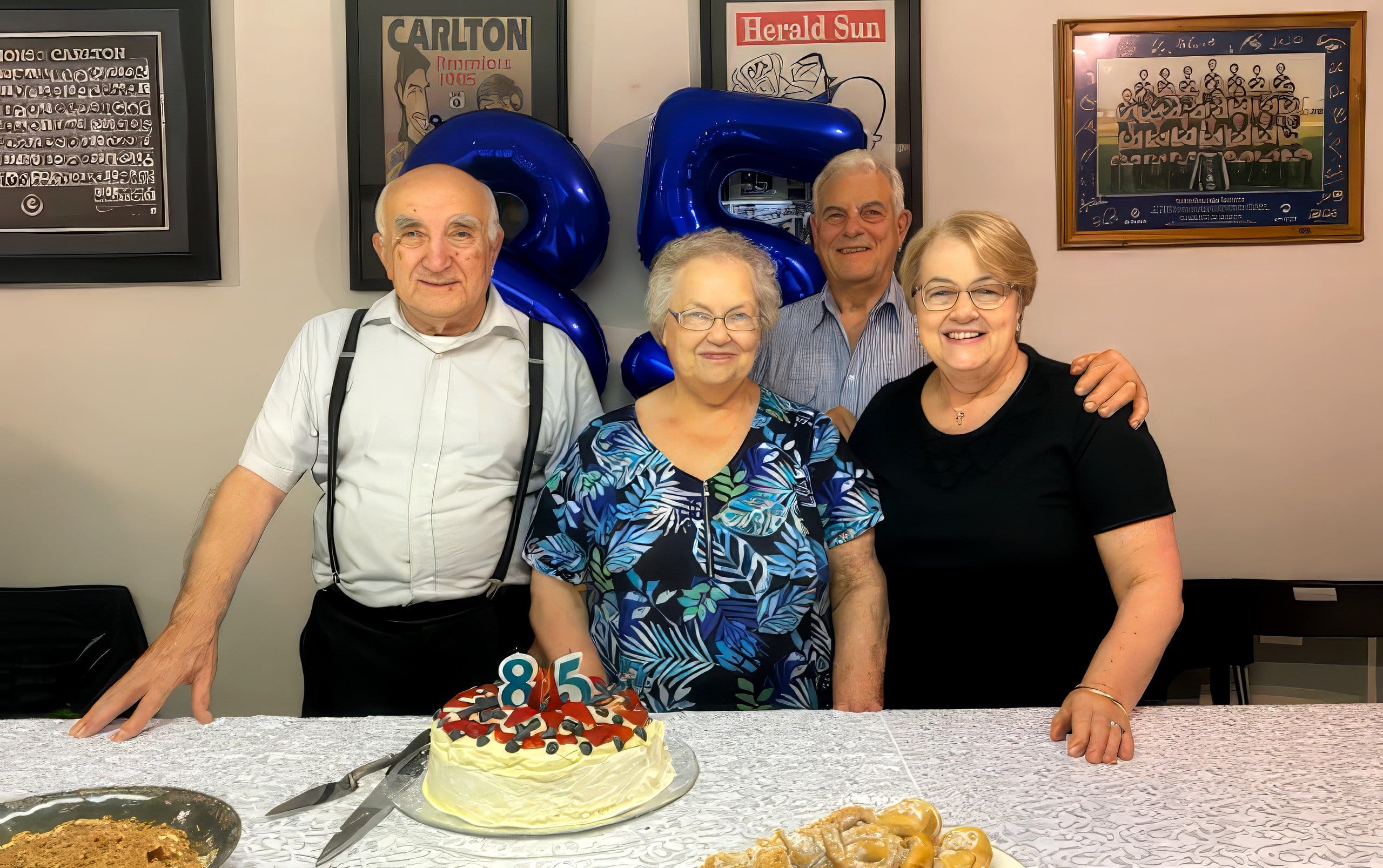 Four older adults standing behind a table with an 85th birthday cake and pastries indoors.