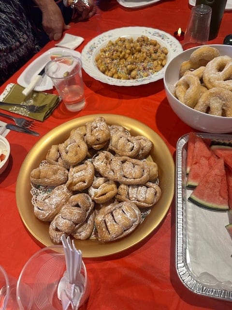 Table with Italian pastries, sugared cookies, fried dough balls, watermelon slices, and plastic cups on a red tablecloth.