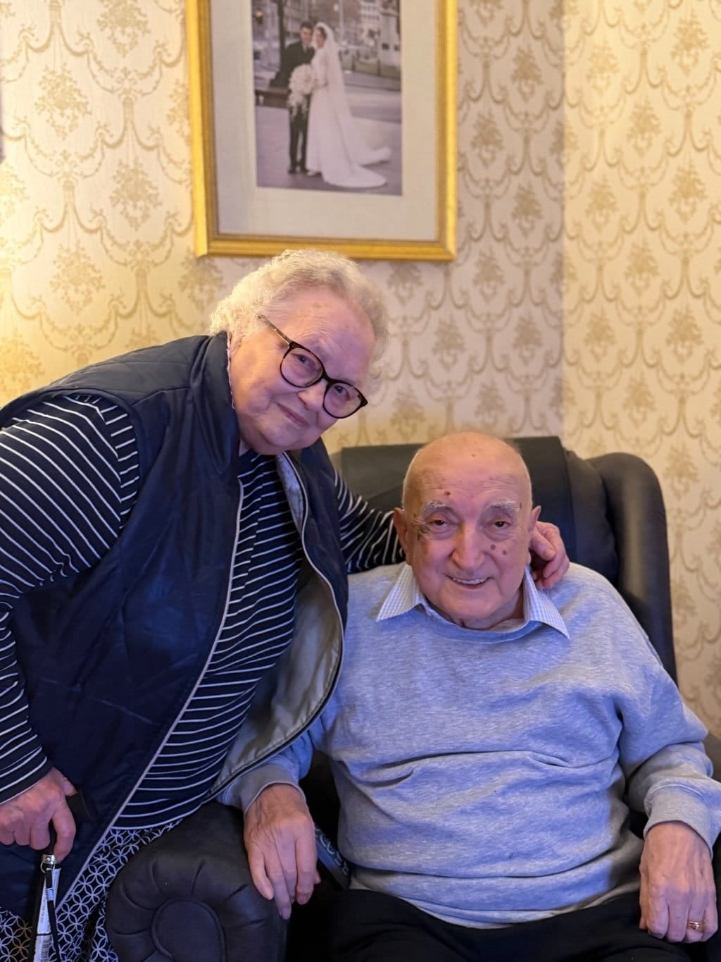 Elderly woman standing beside seated elderly man in a room with patterned wallpaper and a framed wedding photo.